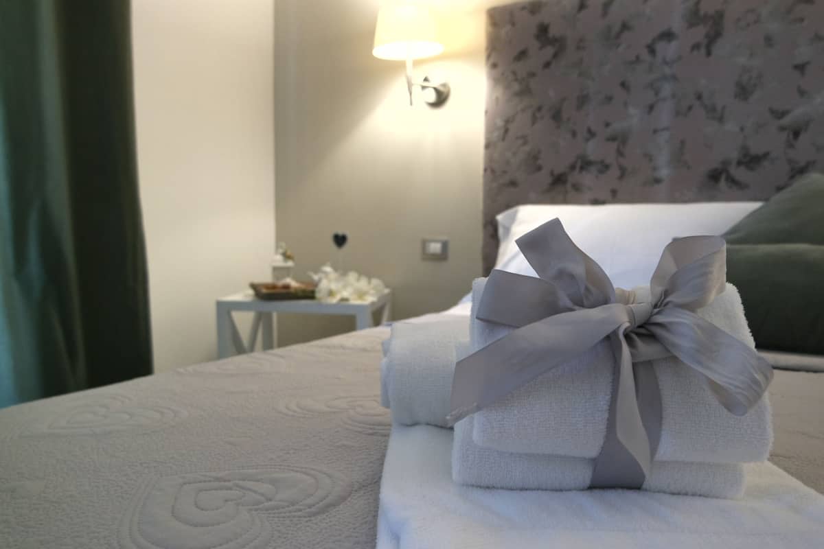 Cama bem arrumada com toalha dobrada e laço, uma pequena mesa com tigela e flores, e uma luminária na parede.