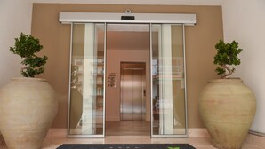 Entrada de un hotel con puerta corredera de vidrio, felpudo con el nombre del hotel y dos grandes macetas a cada lado.