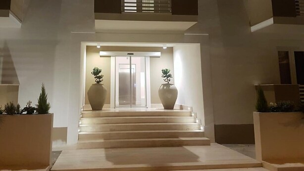 Una entrada moderna a un edificio, con una escalera que lleva a una puerta, dos macetas grandes con árboles y un balcón con barandillas.