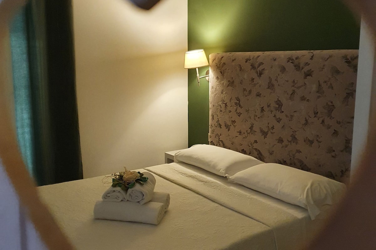Quarto de hotel com um porta-retrato em formato de coração, cama com lençóis brancos, cabeceira estampada com flores, abajur e uma pequena planta na cama.