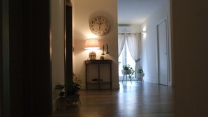 Un pasillo con suelo de madera, un reloj en la pared, una mesa con una lámpara y velas, y plantas en macetas.