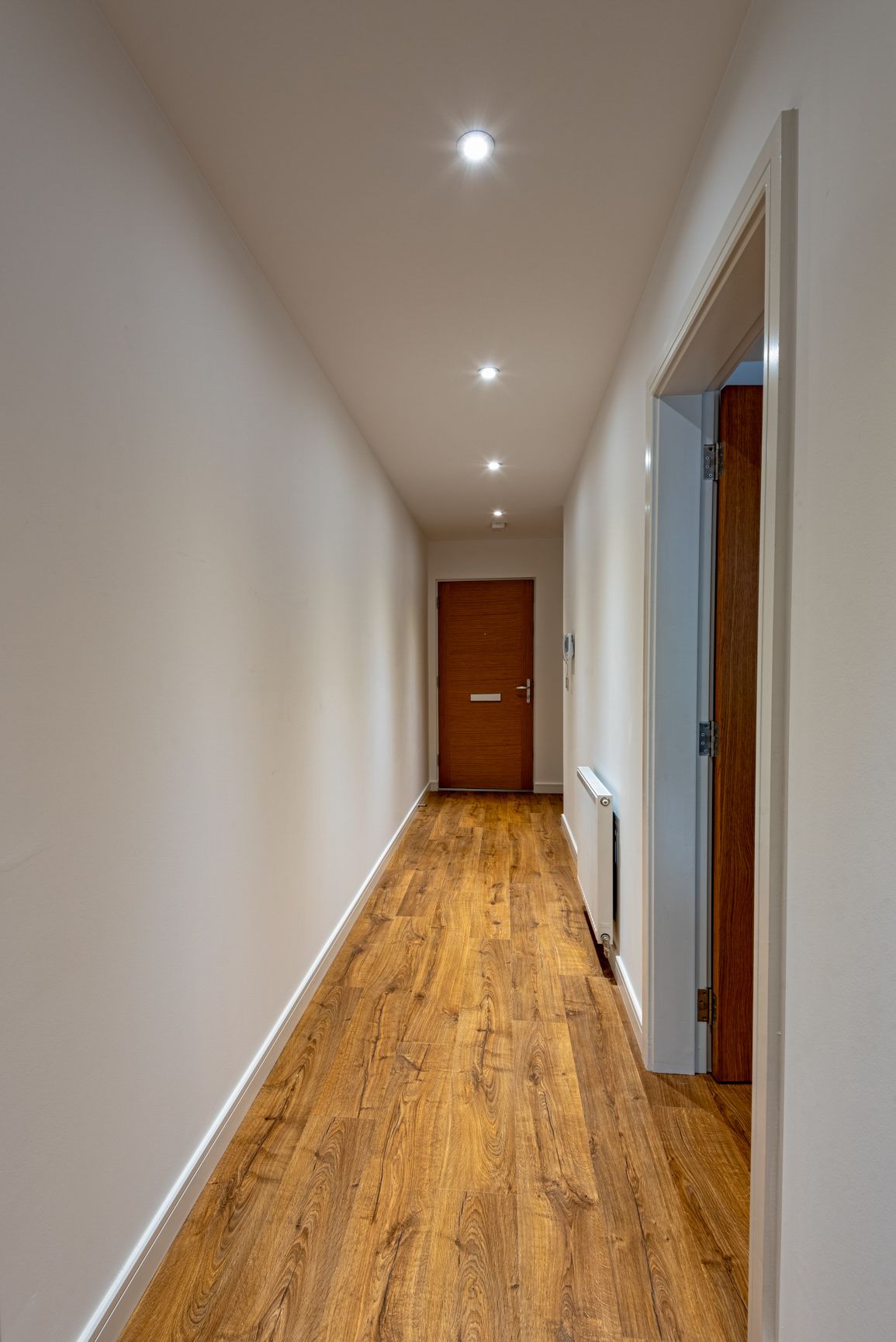 A hallway with wooden flooring, white walls, and recessed ceiling lights.