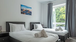 A modern bedroom with two beds, a large window with curtains, a cityscape painting, and a hanging light fixture. - Spacious Modern Apartment (Edinburgh)