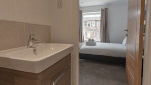 Une salle de bain moderne avec un lavabo et un miroir, une chambre avec un lit et une fenêtre, et une vue sur les bâtiments environnants.