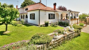 Une maison à deux étages, avec un toit en tuiles, un jardin bordé d’un mur en pierre et agrémenté de sculptures en forme de cygnes, ainsi qu’une pergola couverte de fleurs.