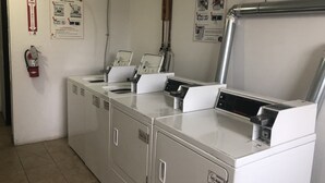 A laundry facility with multiple washing machines and dryers, fire extinguisher, and safety posters on the wall. - Troy Inn and Suites (Troy)