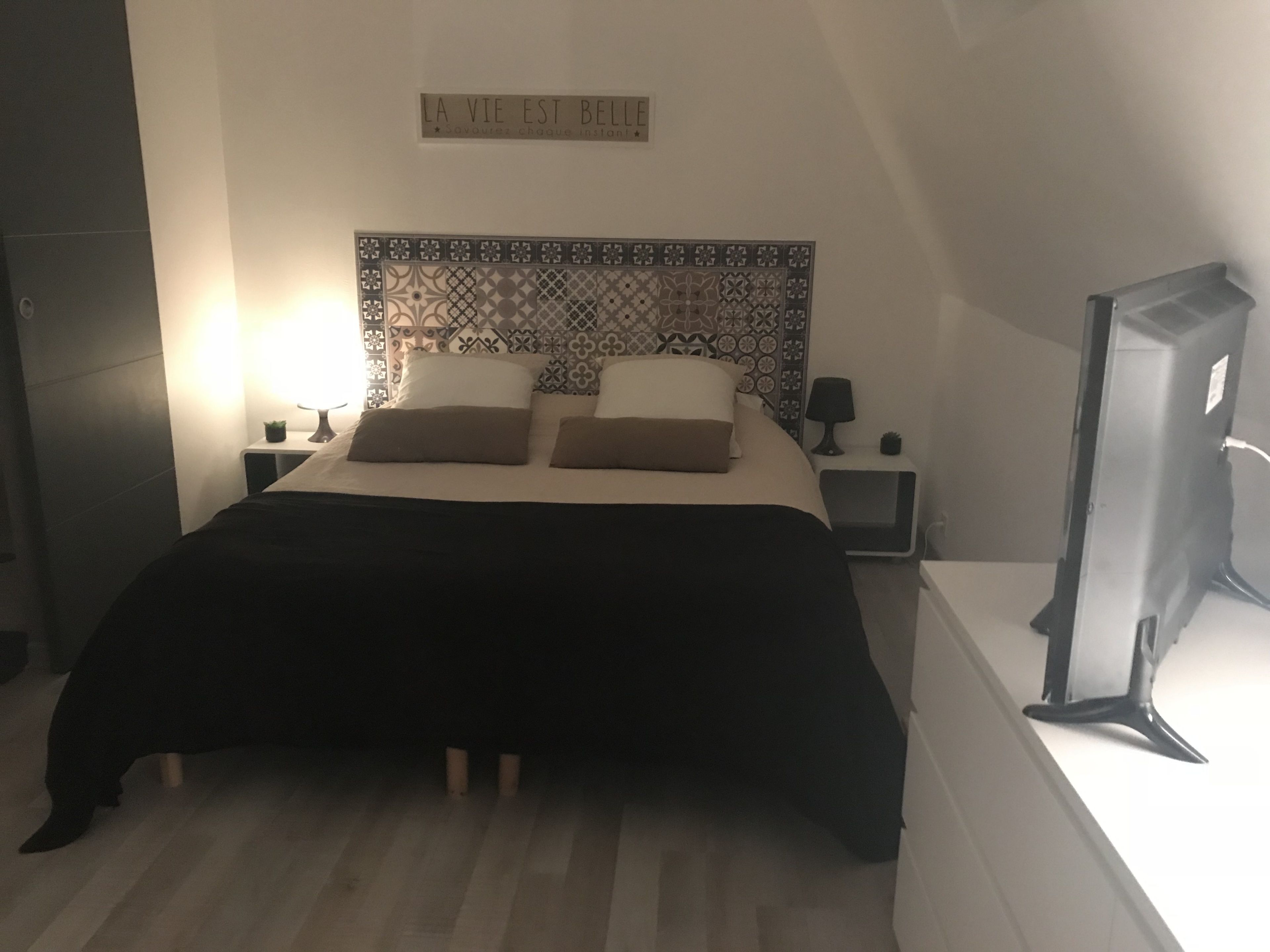Una cama bien hecha con un cabecero estampado, una mesita de noche con lámpara y una señal en la pared.