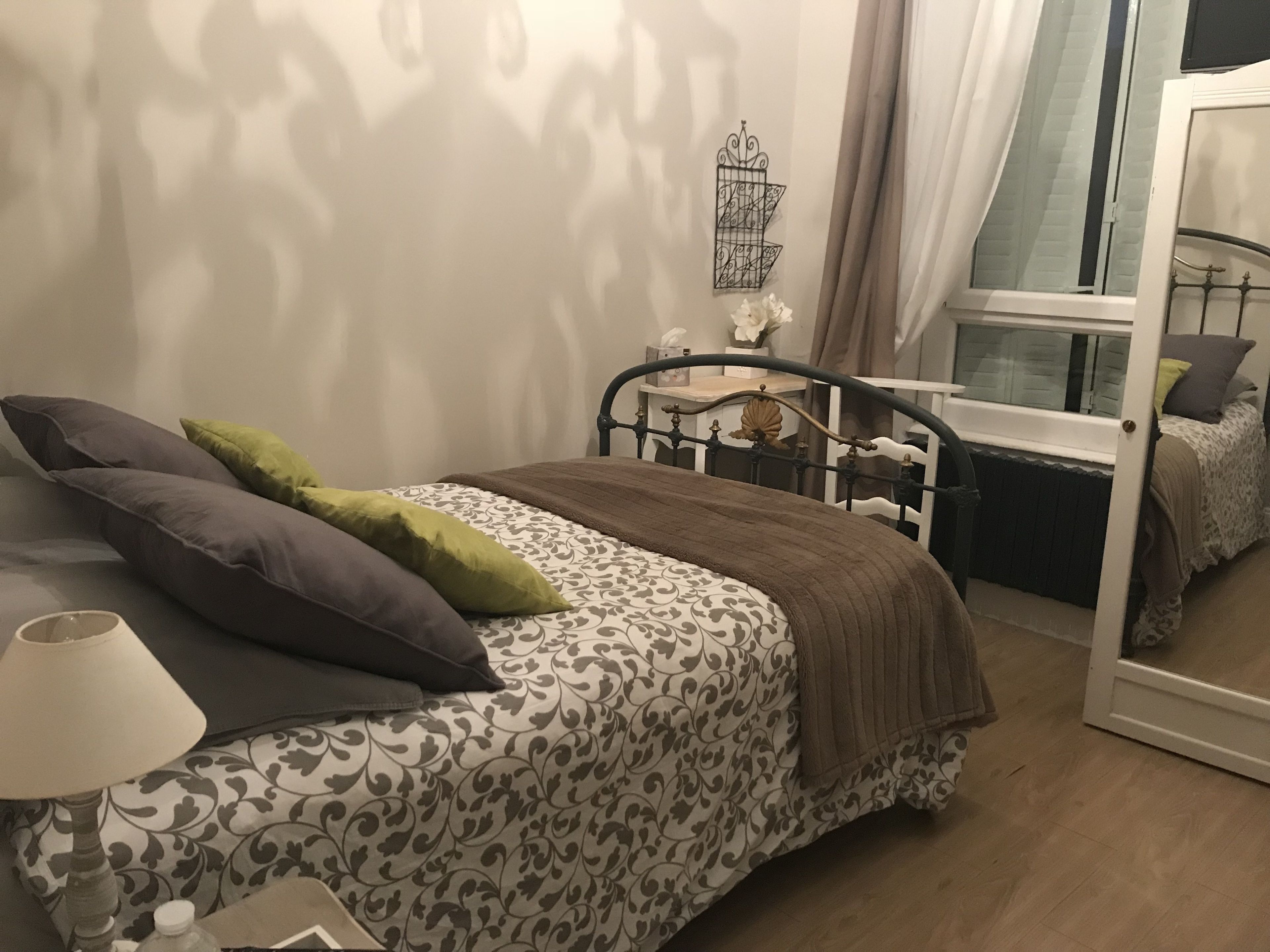Un dormitorio con cama, almohadas, mesita de noche, lámpara, ventana y espejo.
