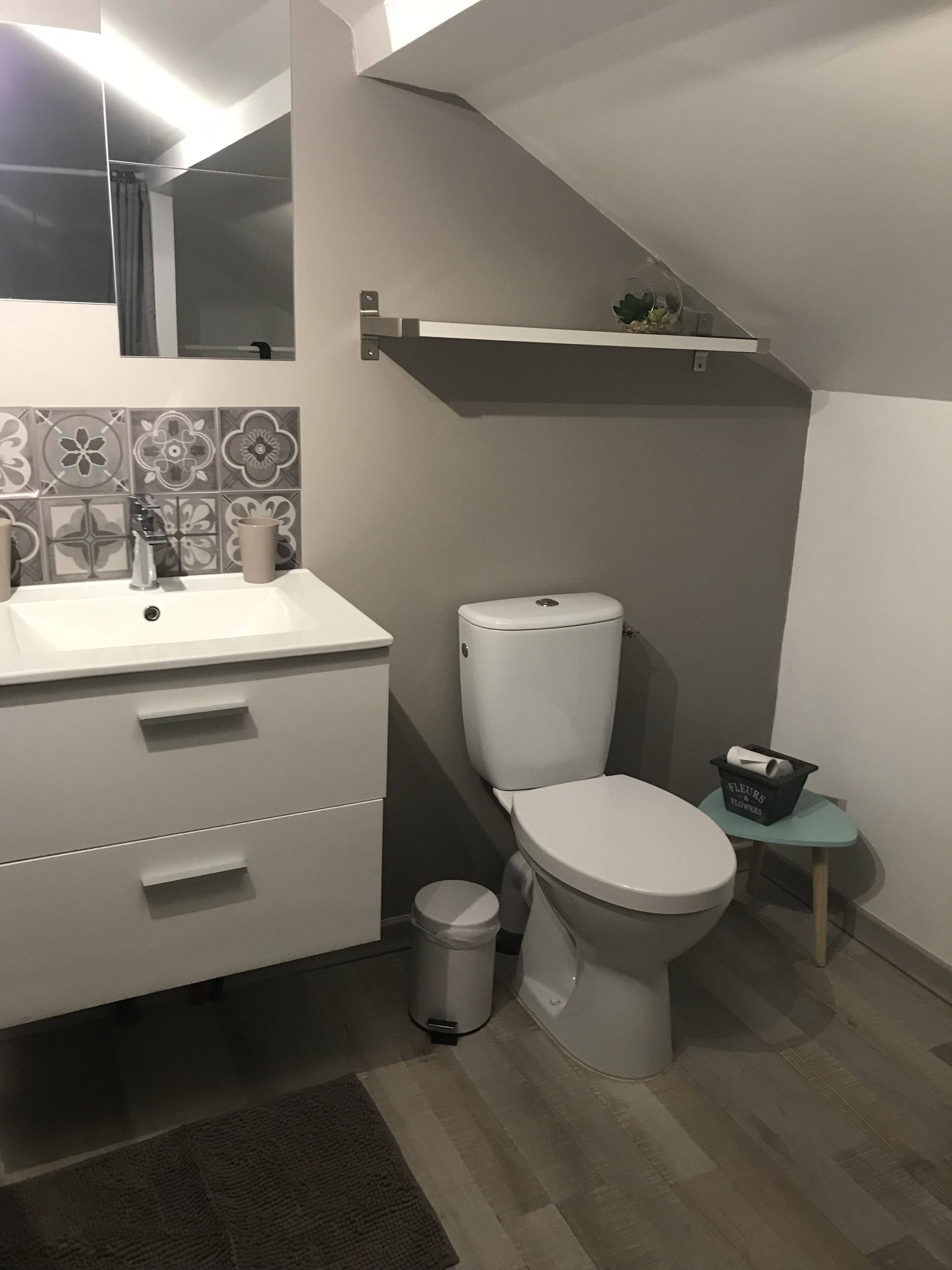 Une salle de bain moderne avec un lavabo, des toilettes et une petite poubelle, le tout de couleur blanche. Il y a une étagère au-dessus des toilettes et un miroir au-dessus du lavabo.