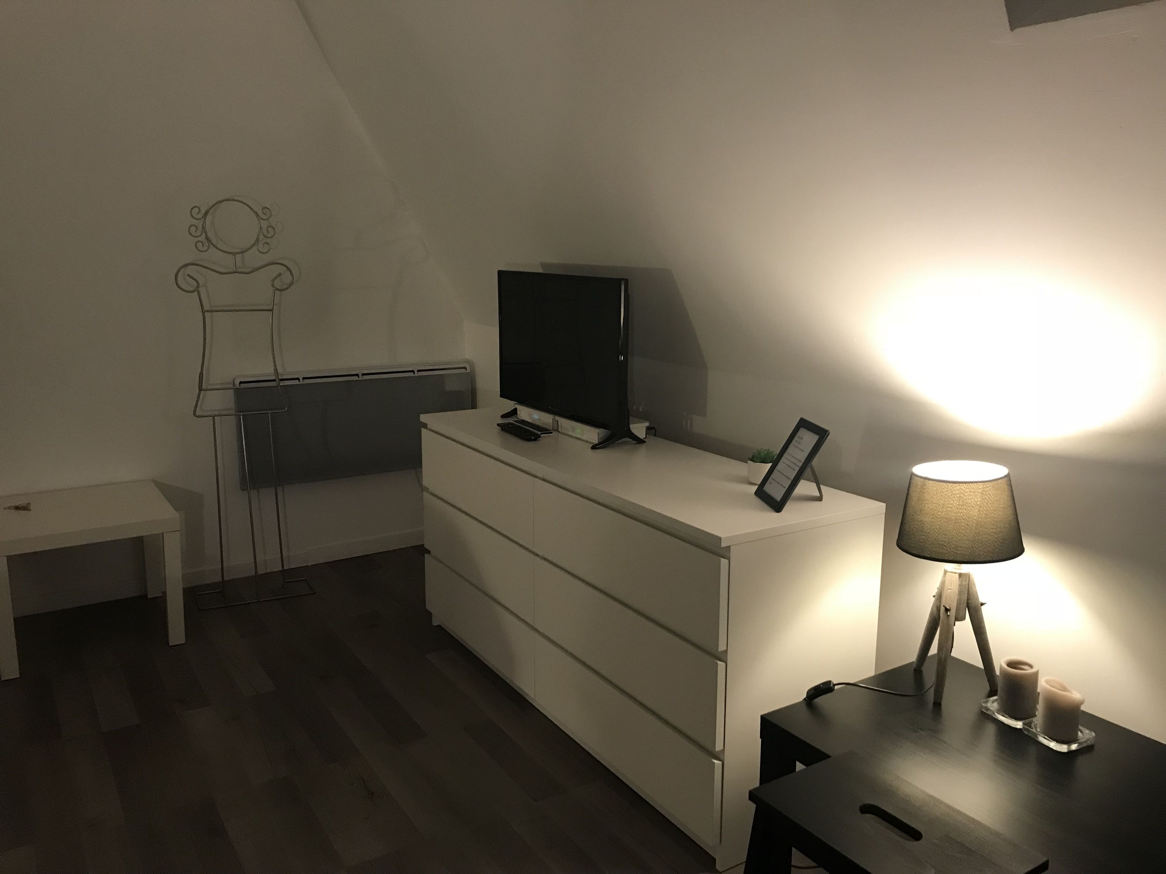 Habitación minimalista con una televisión de pantalla plana sobre un aparador, una mesita con una lámpara y un estante en la pared con una planta.