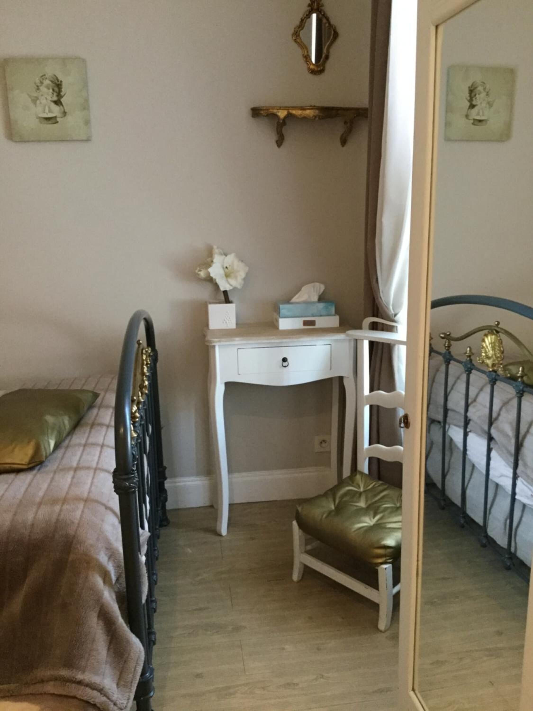 A bedroom with a bed, a desk, a chair, a mirror, and a shelf with a vase of flowers.