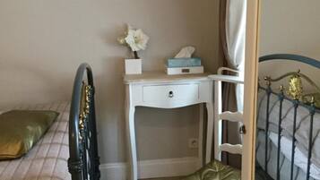 A bedroom with a bed, a desk, a chair, a mirror, and a shelf with a vase of flowers.