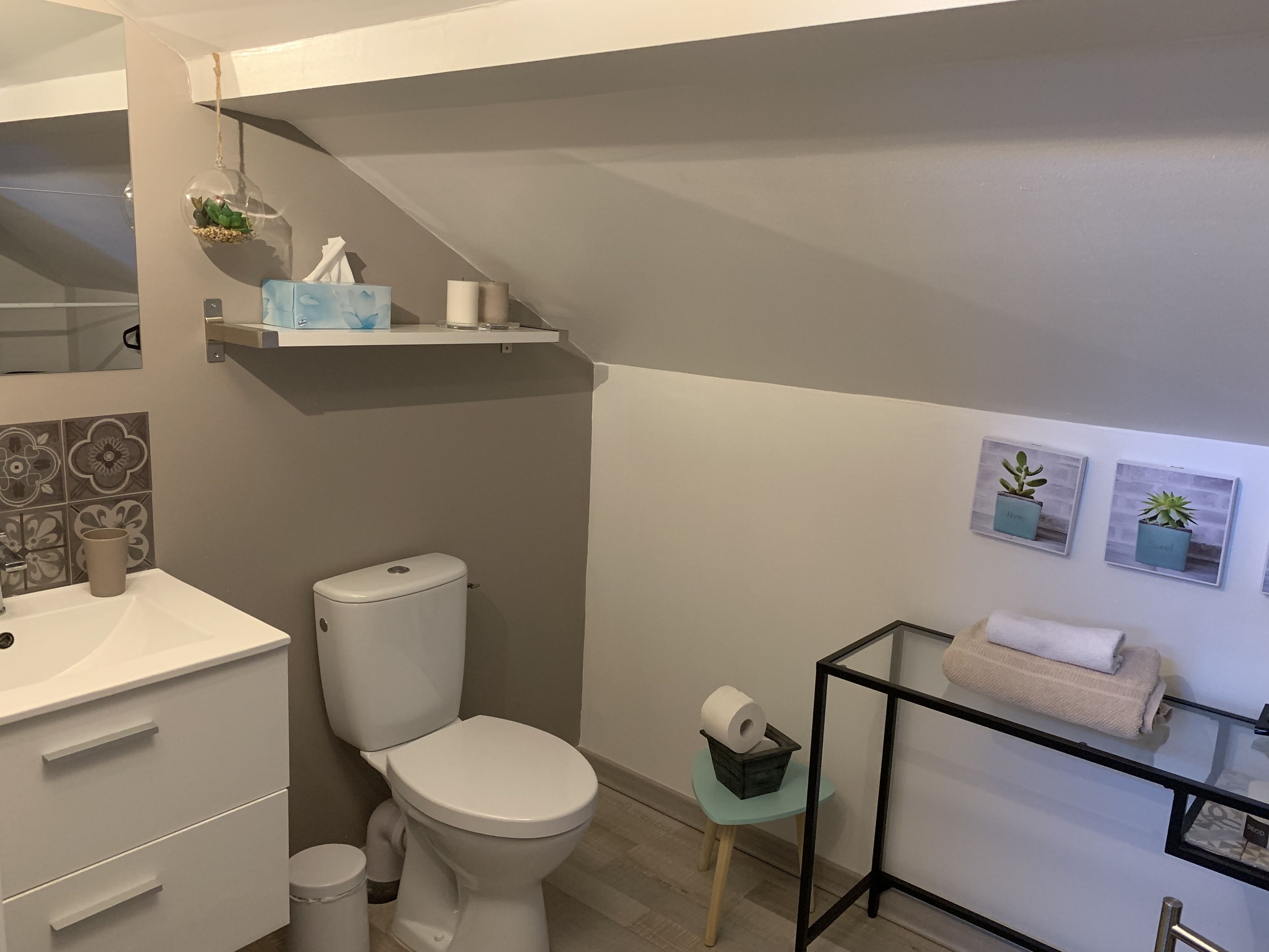 A modern bathroom with a toilet, sink, and storage cabinets. There is a shelf with toiletries and a small plant.