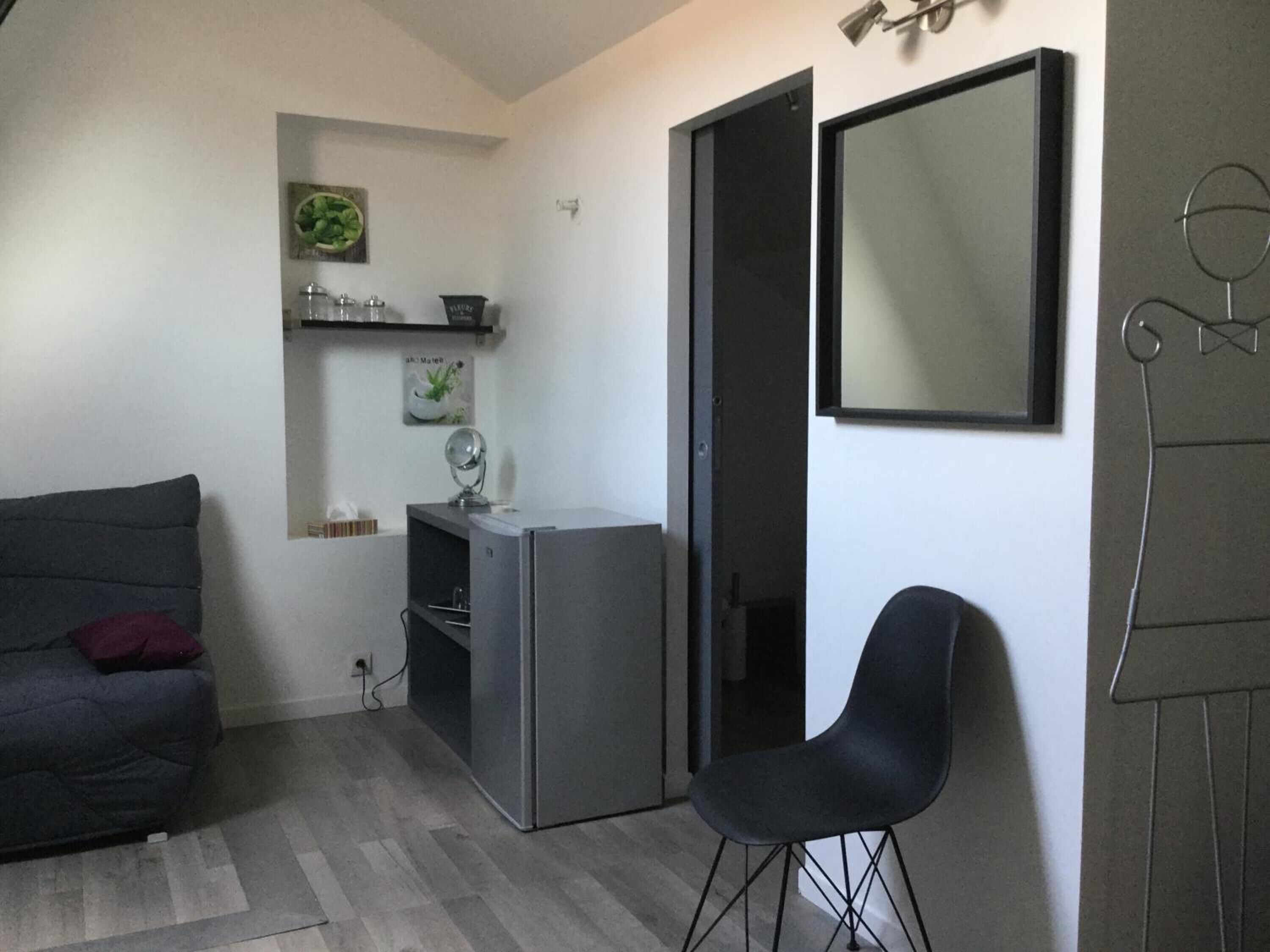 Un espacio habitable pequeño con un sofá gris, un escritorio con lámpara, una silla y un espejo grande en la pared.
