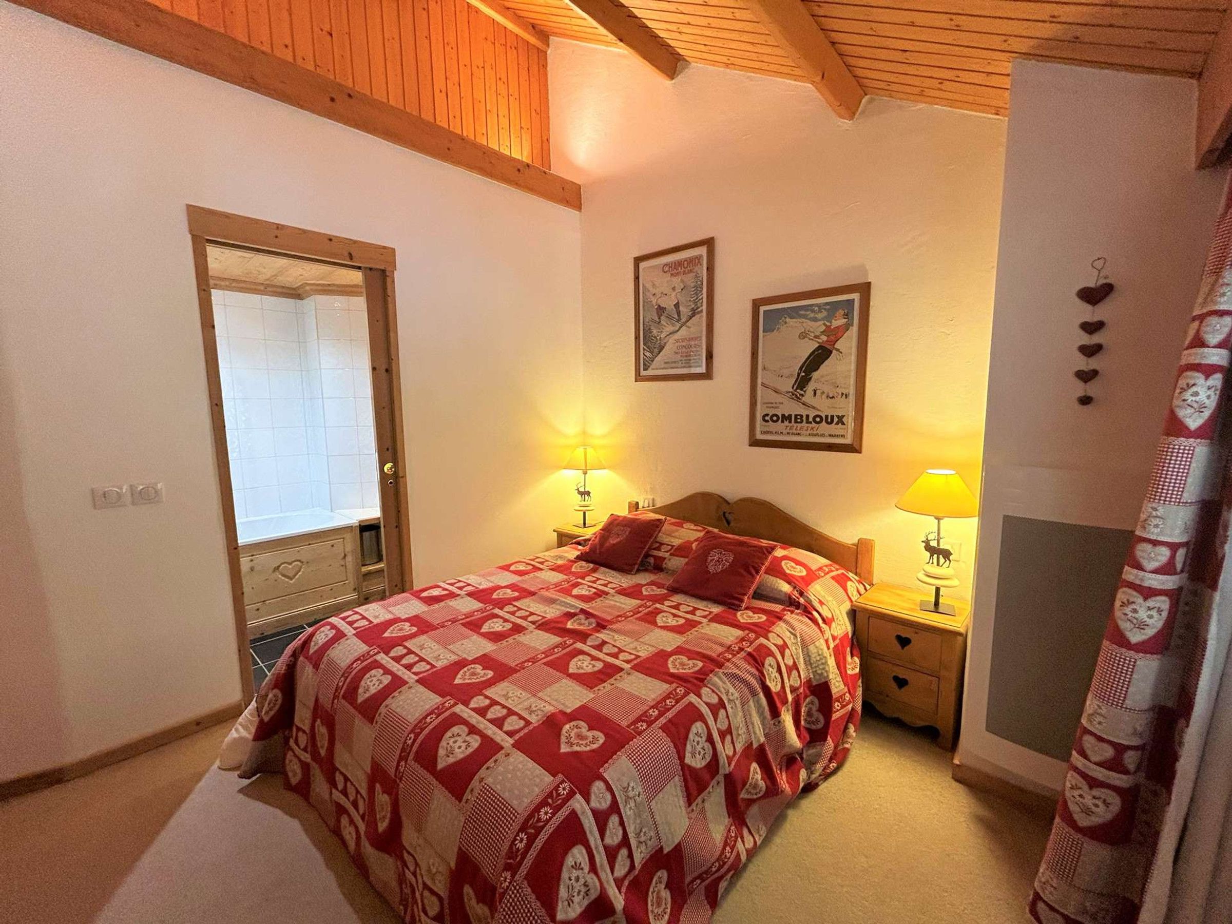 A bedroom with a wooden ceiling, a bed with a red and white quilt, two bedside lamps, a mirror, and a door.