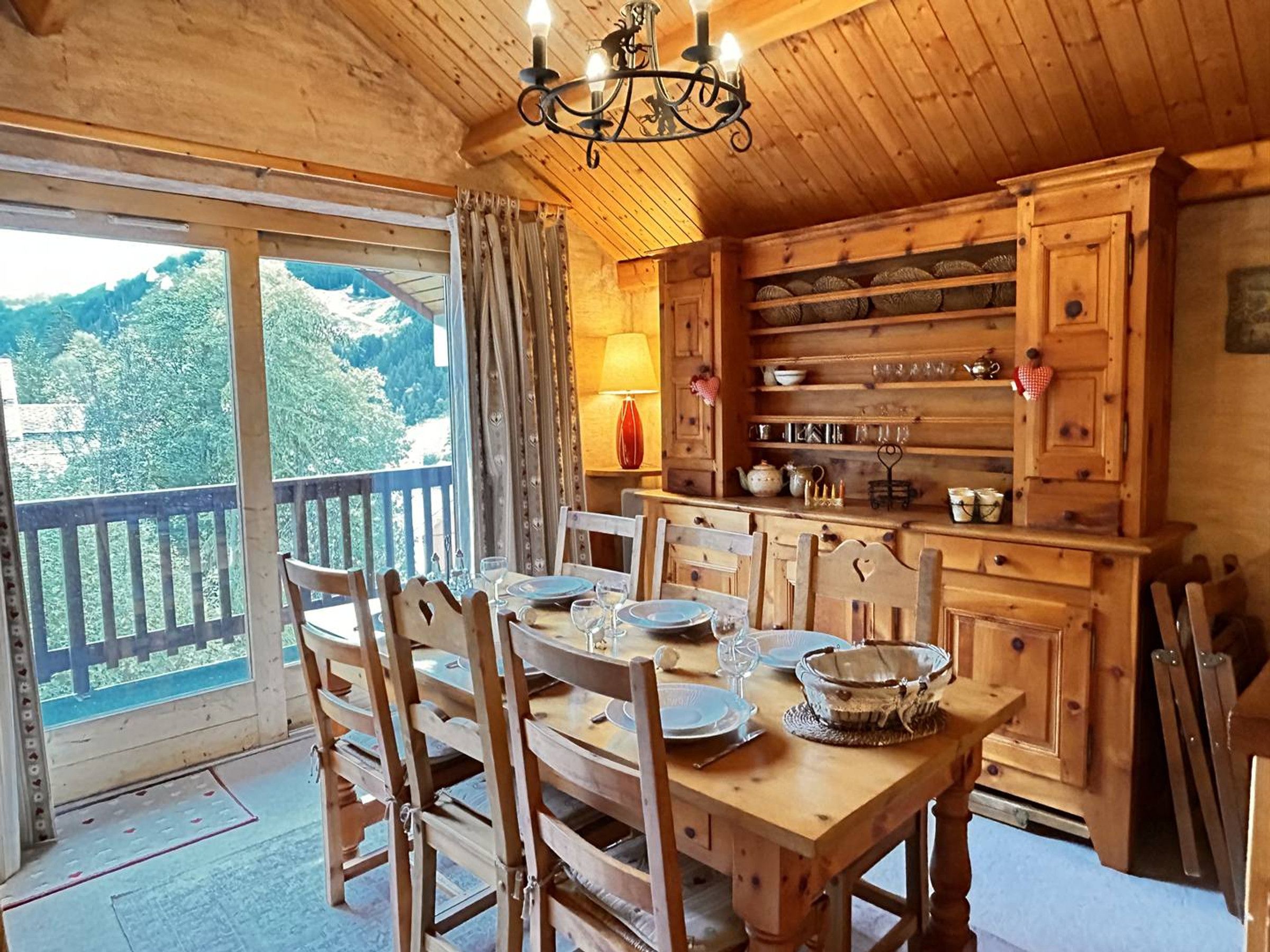 Comedor de madera con una mesa dispuesta para cuatro, sillas, una lámpara de araña y vistas al exterior.