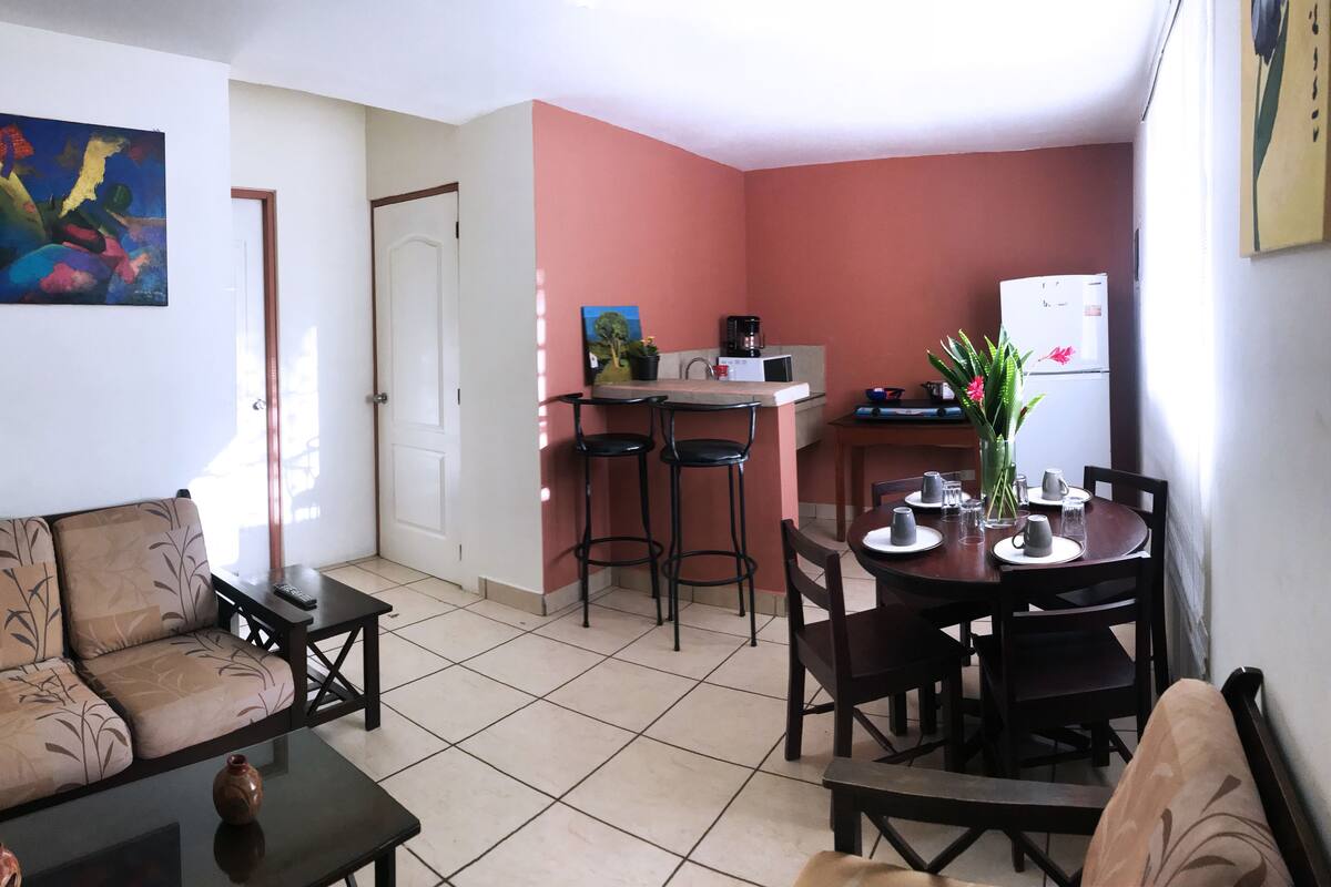 A compact living space with a sofa, a dining table with chairs, a kitchen area with a refrigerator, and a painting on the wall.
