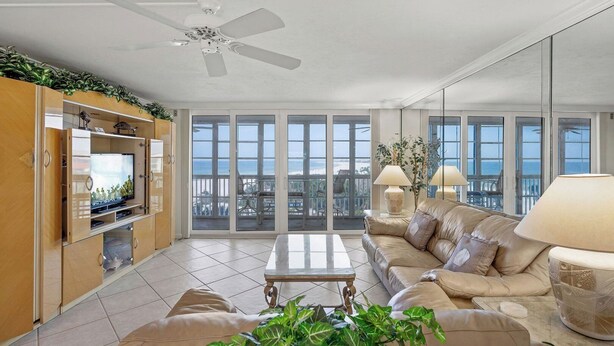 A spacious living room with a large TV, comfortable seating, a ceiling fan, and a balcony with a view. - Crystal Sands Tower Unit 509 - Beachfront (SARASOTA)
