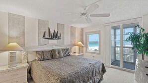 A bedroom with a large bed, two bedside tables, a ceiling fan, a window with a view of the beach, and a potted plant. - Crystal Sands Tower Unit 509 - Beachfront (SARASOTA)