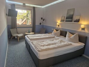 Ein Hotelzimmer mit zwei Betten, einem Schreibtisch, einem Stuhl, einem Fernseher und einem Fenster mit Blick auf ein Gebäude und Bäume.