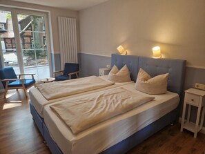 Ein Doppelbett mit zwei Kissen, ein Sessel, ein Nachttisch und ein Fenster mit Jalousien.
