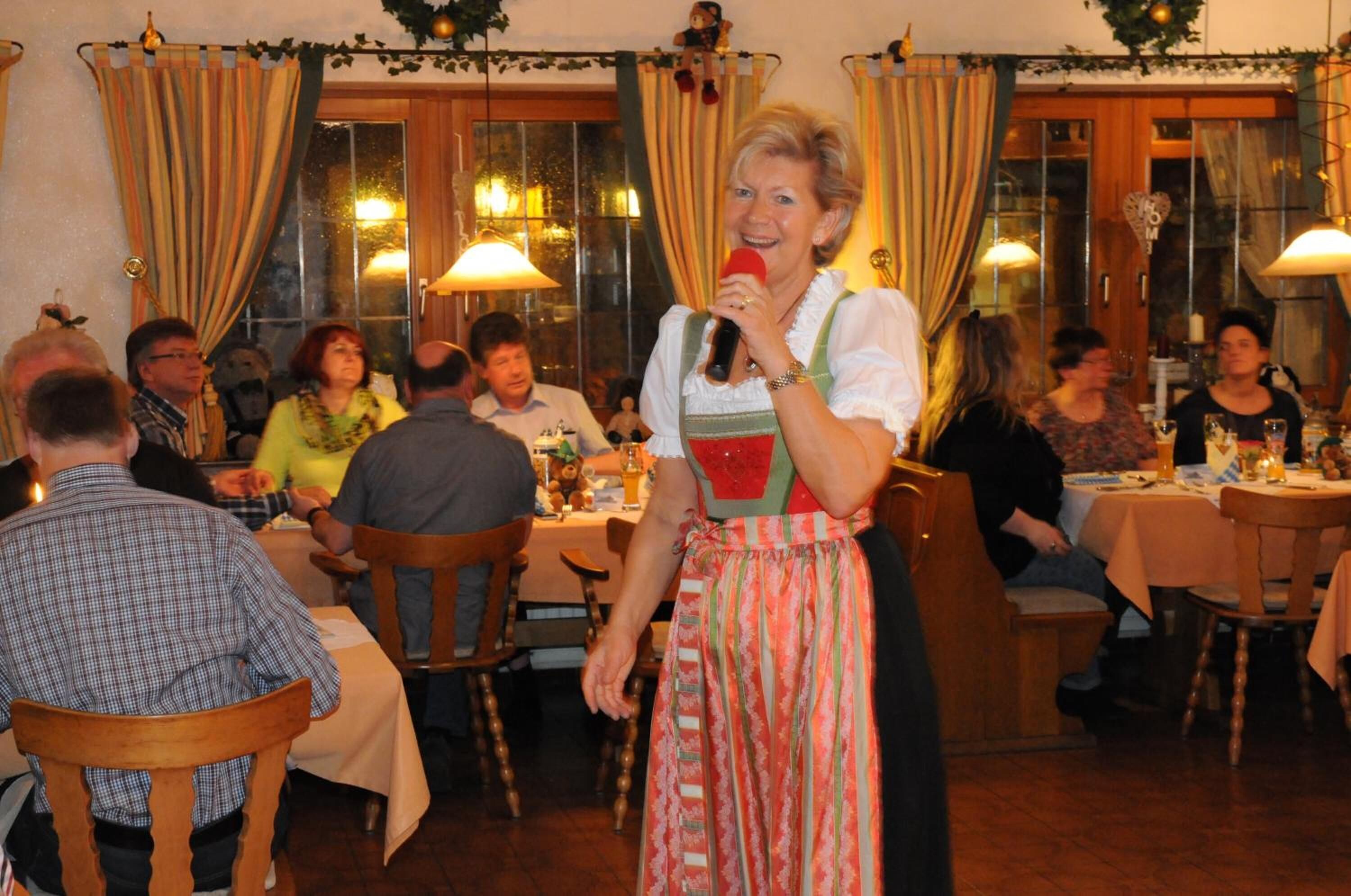 A person speaking at a table in a room with wooden furniture and curtains.