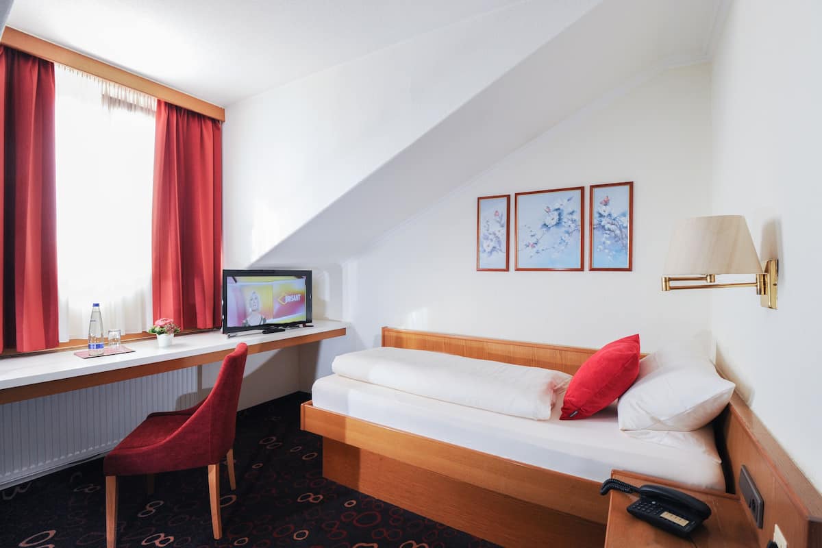 Habitación de hotel con una cama, un escritorio con computadora, una silla, un teléfono y una ventana con cortinas.
