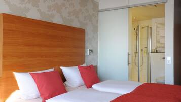 Habitación de hotel con una cama grande, cabecero de madera y zona de ducha visible a través de una puerta de cristal.