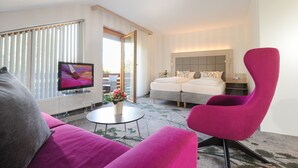 A modern hotel room with a large flat-screen TV, a pink armchair, a sofa, a small round table with a vase of flowers, and a bed with a headboard.