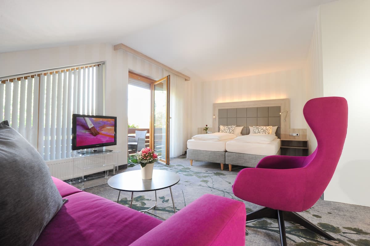 Una habitación de hotel moderna con un televisor de pantalla plana grande, un sillón rosa, un sofá, una pequeña mesa redonda con un jarrón de flores y una cama con cabecera.
