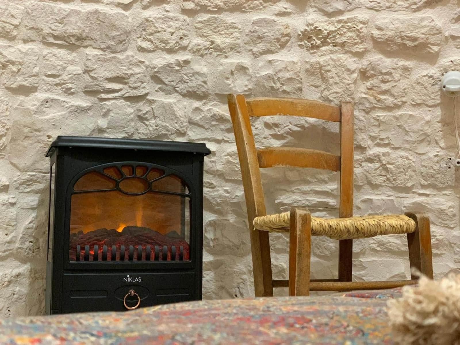 A wood-burning stove with a visible flame, a wooden chair with a woven seat, and a textured stone wall.