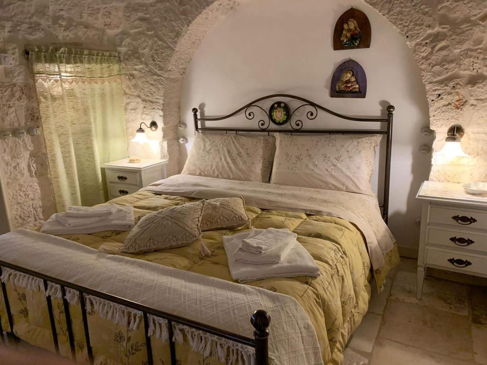 Une chambre avec un mur en pierre, un lit avec une tête de lit, deux tables de chevet avec des lampes et une petite statue religieuse placée au-dessus du lit.