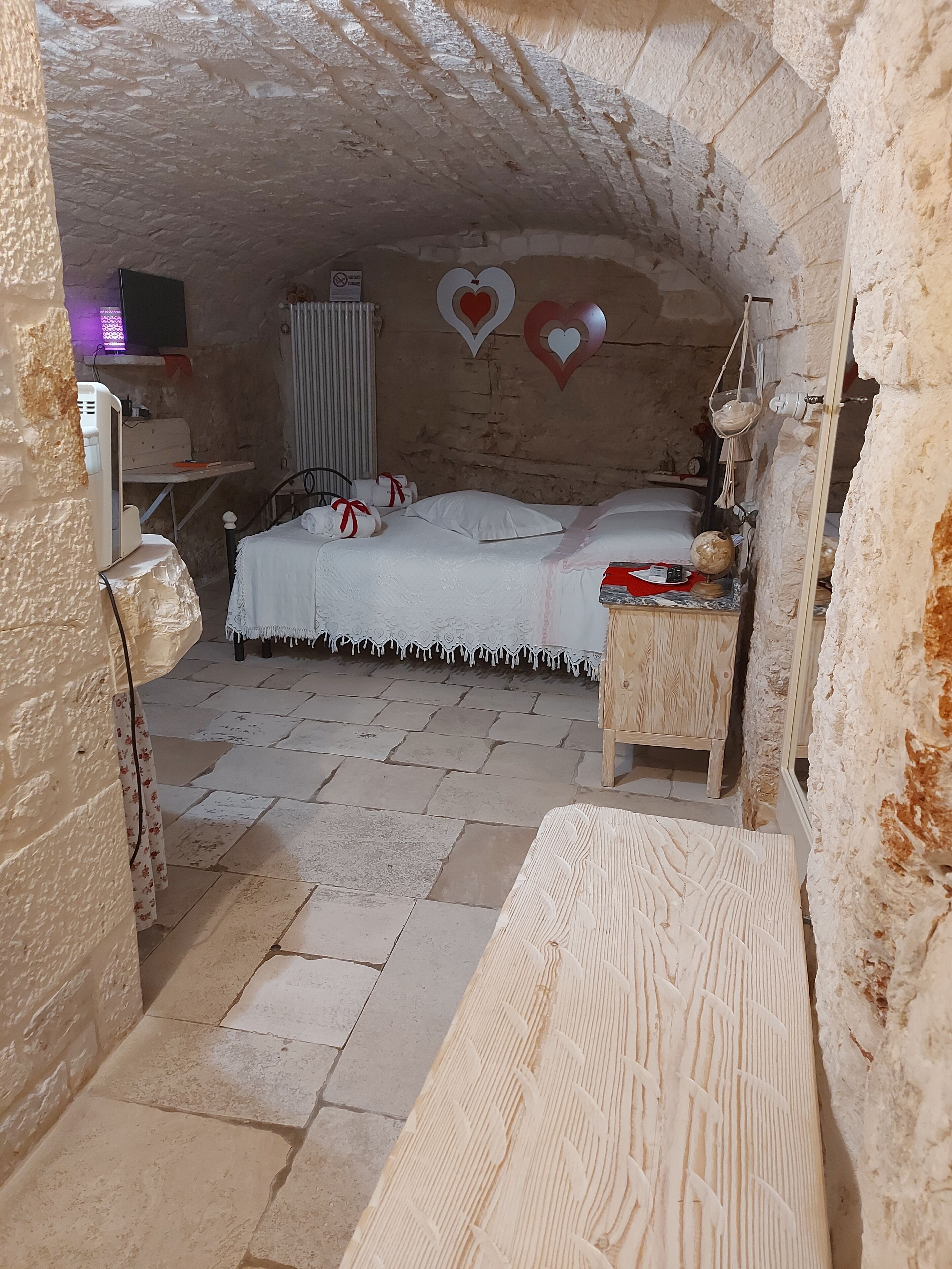 A cozy, rustic room with a stone floor, a bed with white linens, a bedside table with a globe and books, and heart-shaped decorations on the wall.