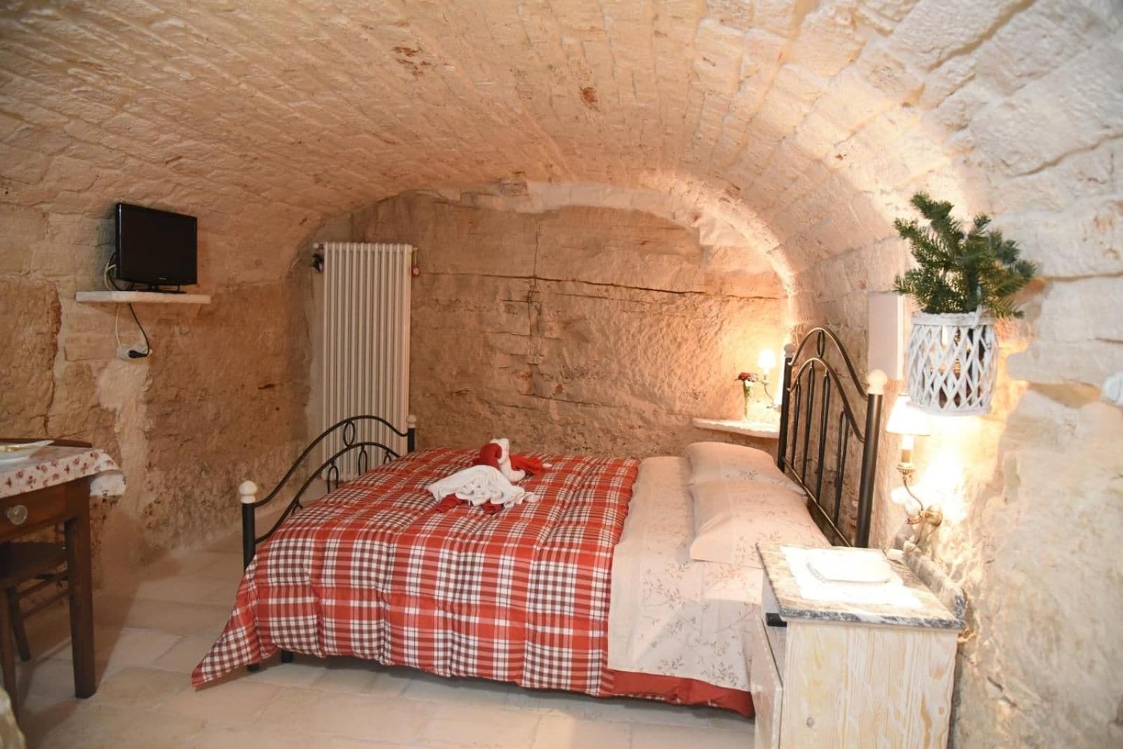 Ein Schlafzimmer mit Stehwand, einem Bett mit rot-weiß kariertem Überwurf, einem Nachttisch mit Lampe und einem kleinen Regal mit einer Pflanze.