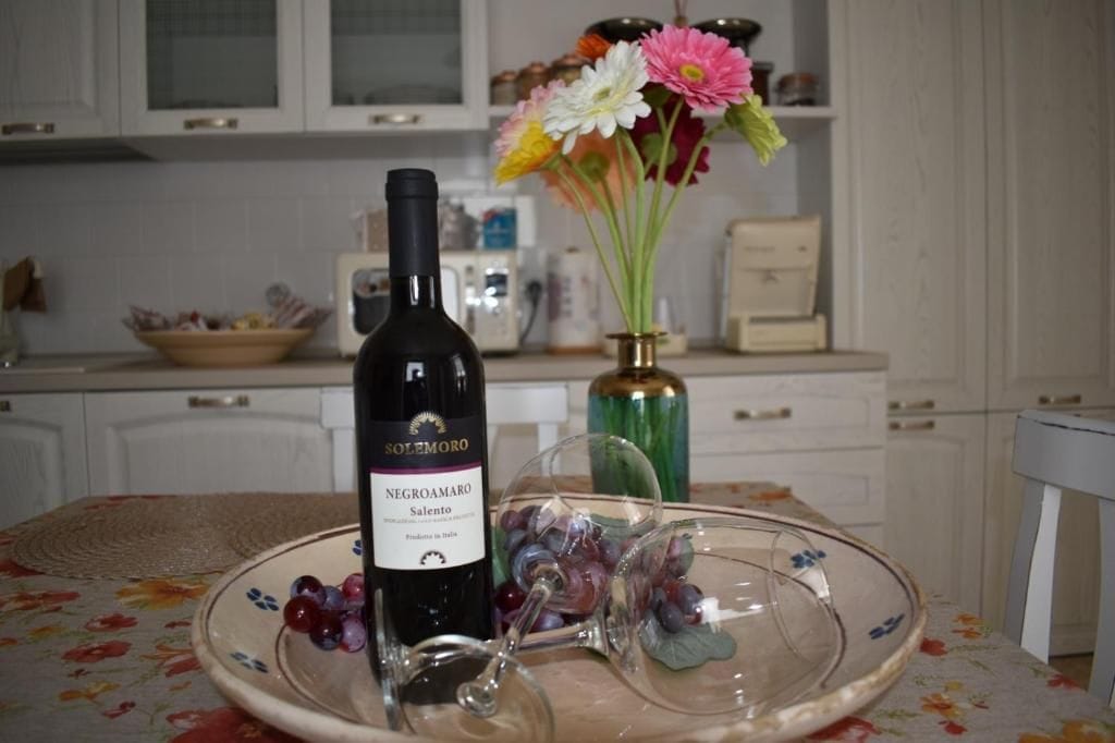 Una bottiglia di vino Negroamaro, un bicchiere e un piatto con acini d'uva su un tavolo in una cucina.