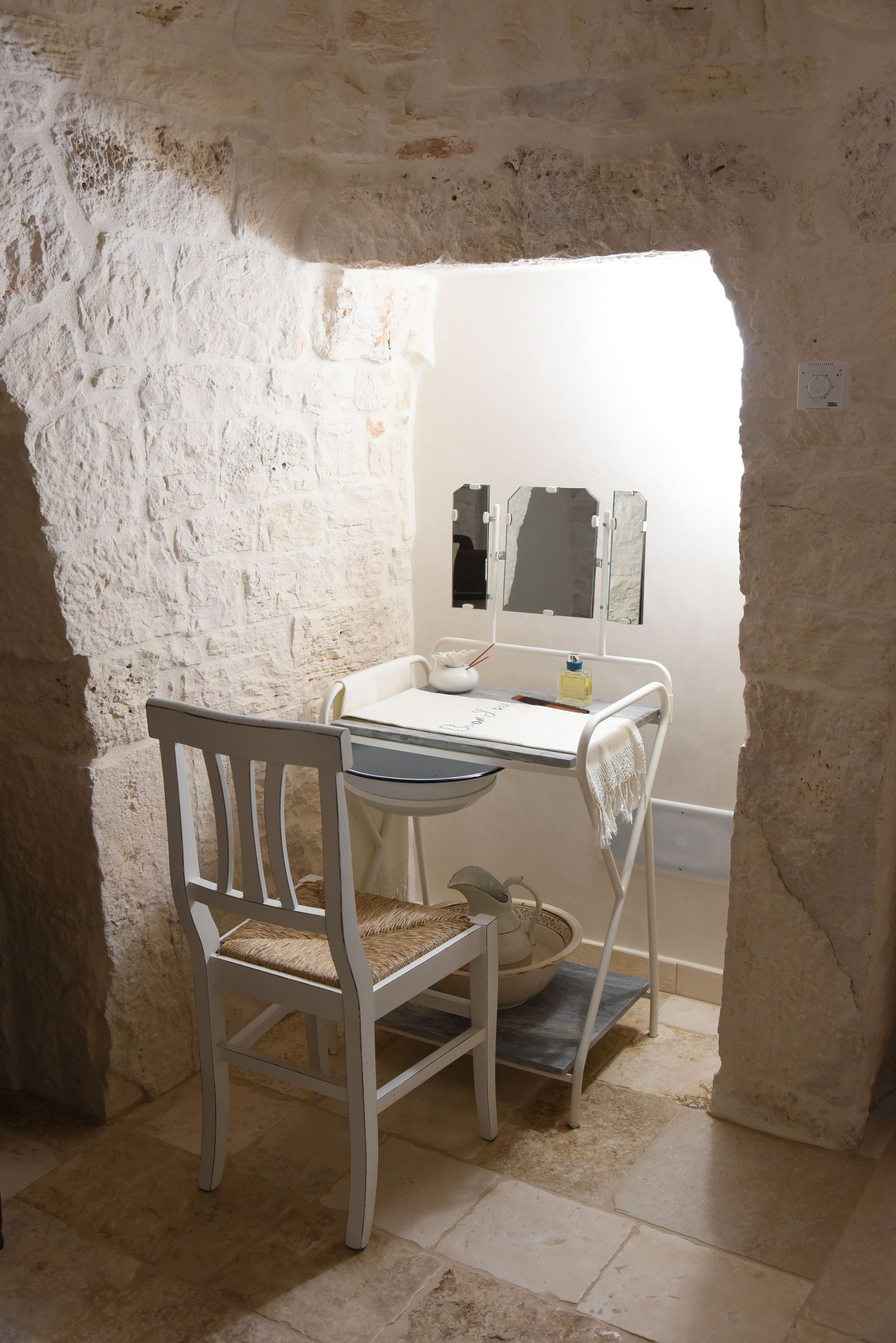 A small, rustic room with a white table and chair, a mirror on the wall, and a stone wall.