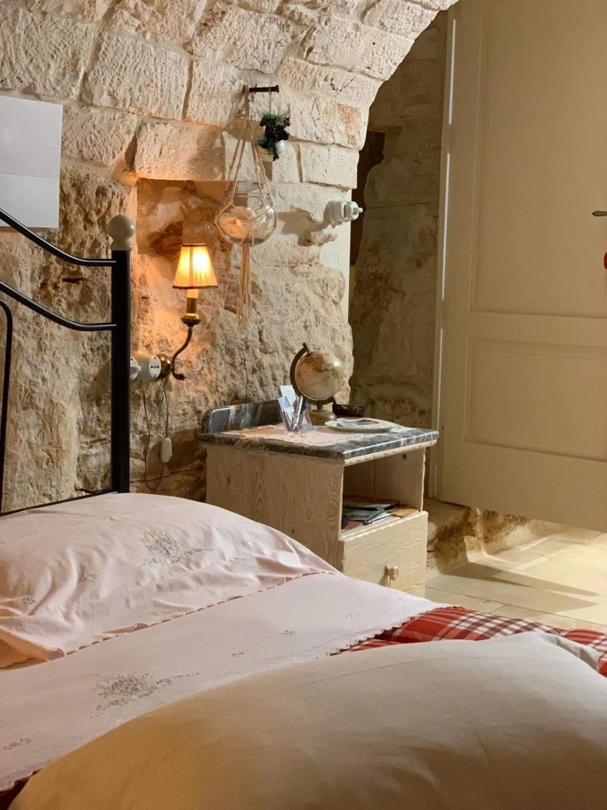 Une chambre avec un mur en pierre, une lampe de chevet, une petite table en bois et un lit recouvert d’une couverture blanche.