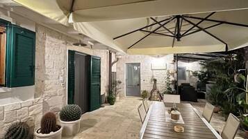 Una zona patio esterna con un lungo tavolo di legno e sedie, circondata da cactus e piante in vaso, sotto un ombrellone grande.