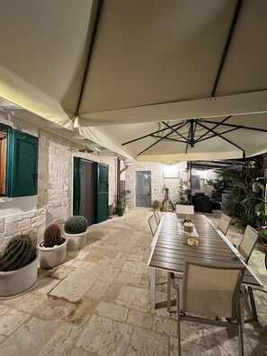 An outdoor patio area with a long wooden table and chairs, surrounded by potted cacti and plants, under a large umbrella. - Trulli Occhi di Pietra (Alberobello)