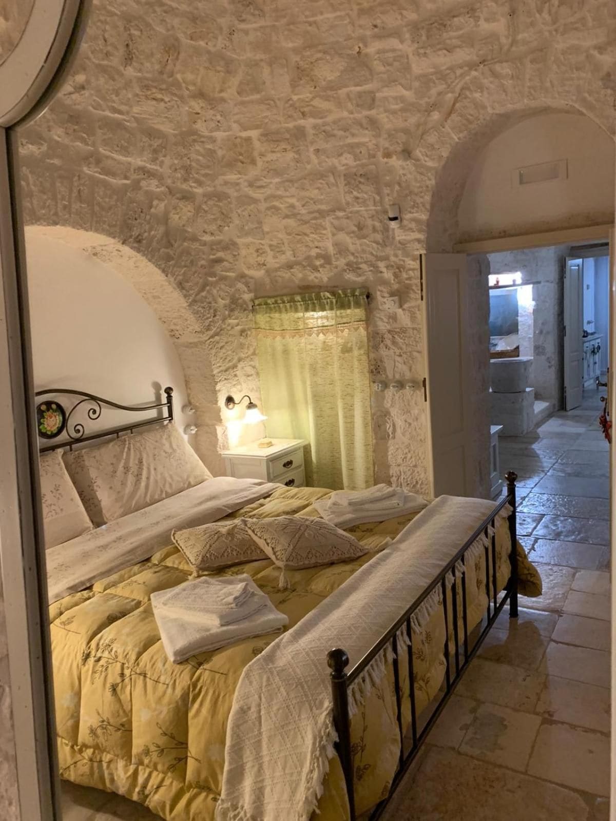 A bedroom with a stone wall, a bed with a metal frame, a nightstand with a lamp, and a window with a curtain.