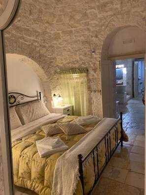 Un dormitori amb paret de pedra, un llit amb estructura metàl·lica, una tauleta de nit amb una làmpada i una finestra amb cortina.