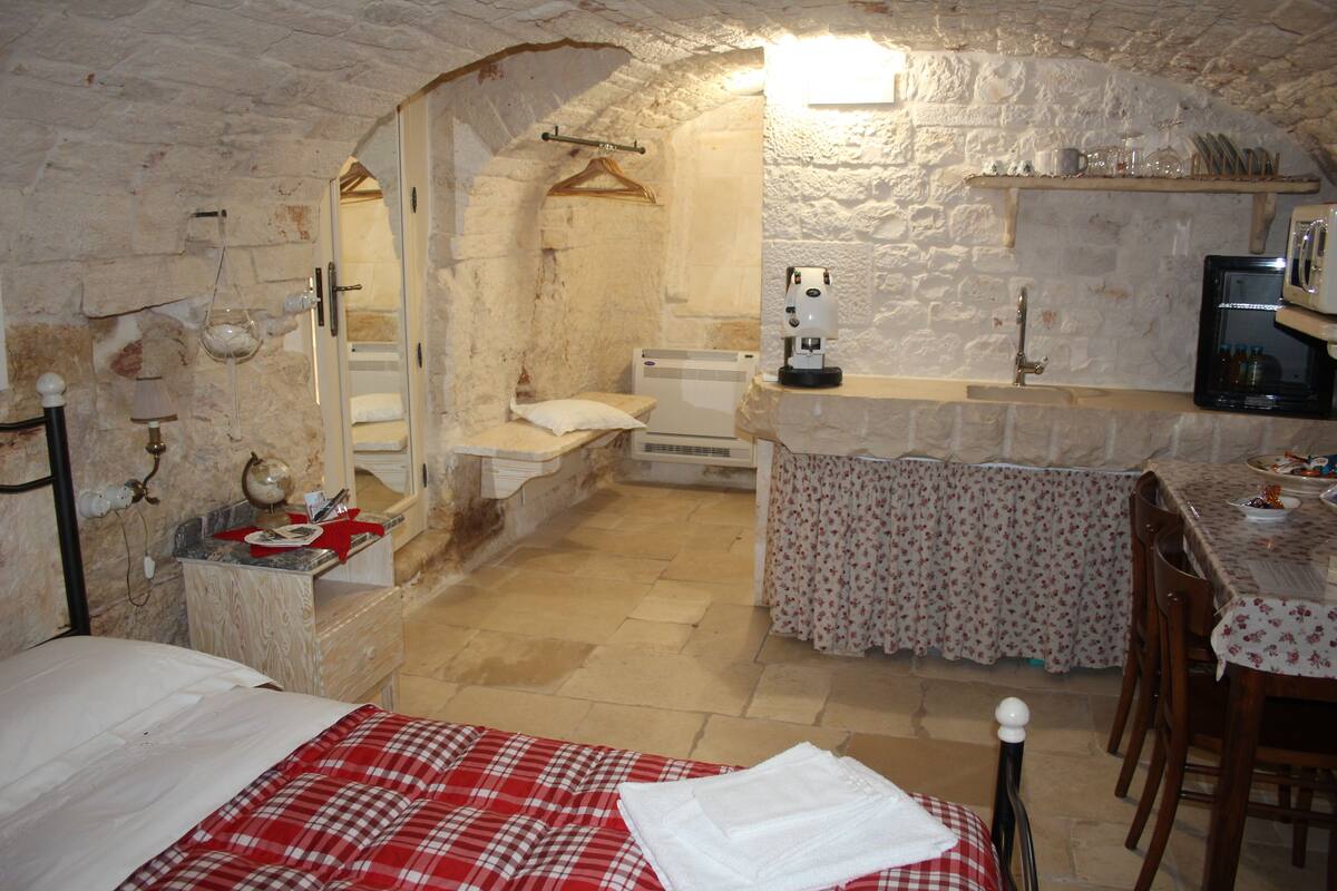 A bedroom with a bed, a small table with a chair, a kitchenette with a coffee machine, and a bathroom with a mirror and sink.