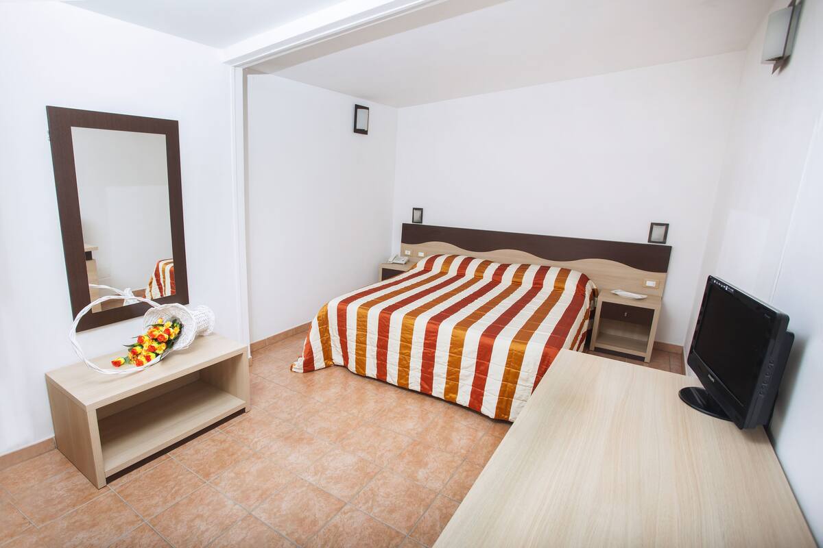A bedroom with a bed, a TV, a bedside table, a mirror, and a console table with a basket of flowers.