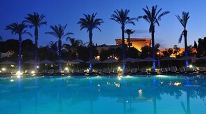 A pool area with lounge chairs and umbrellas, palm trees, and a building in the background. - Green Paradise Alimini Resort (Otranto)