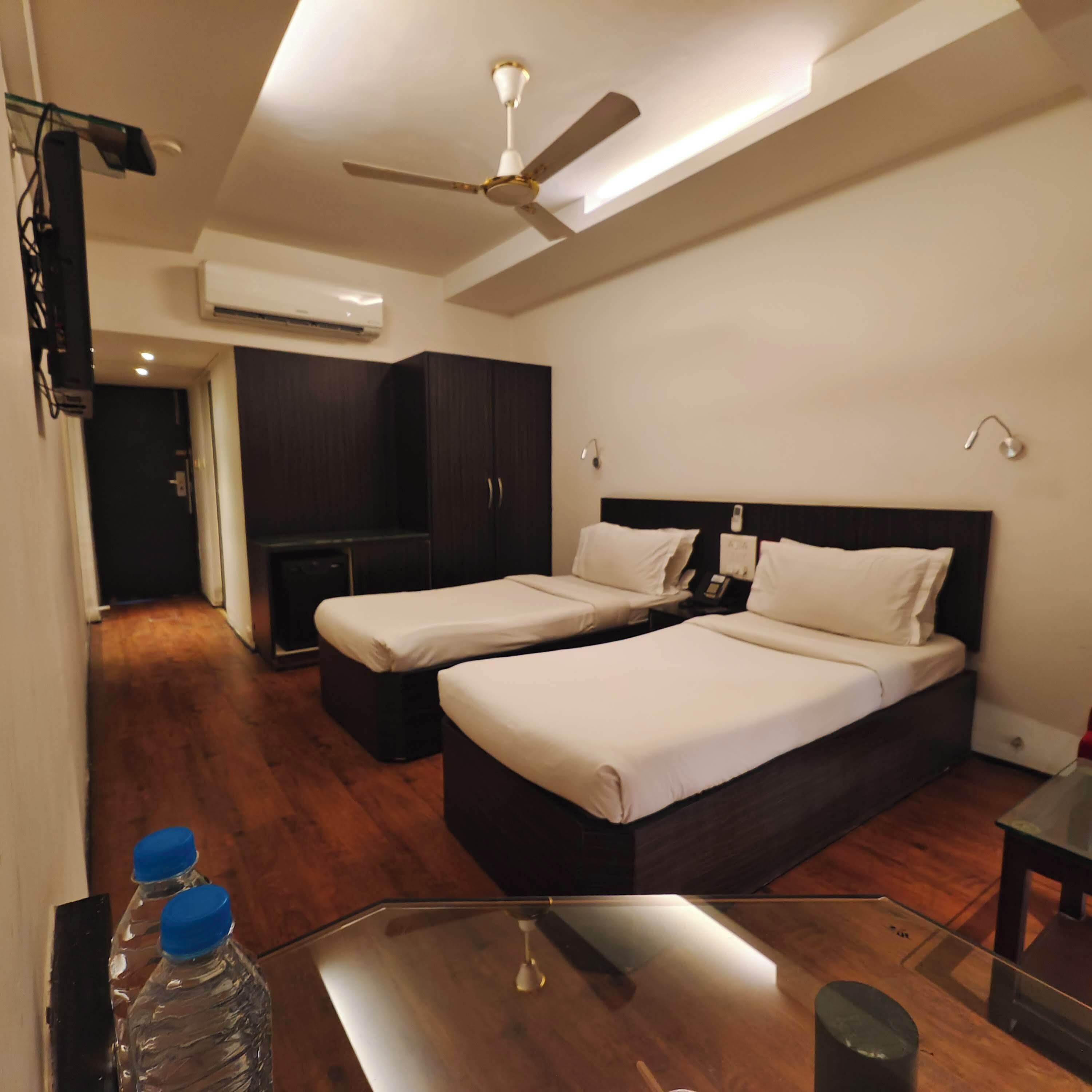 Habitación de hotel con dos camas, ventilador de techo, unidad de aire acondicionado, suelo de madera y mesa de vidrio con botellas de agua.