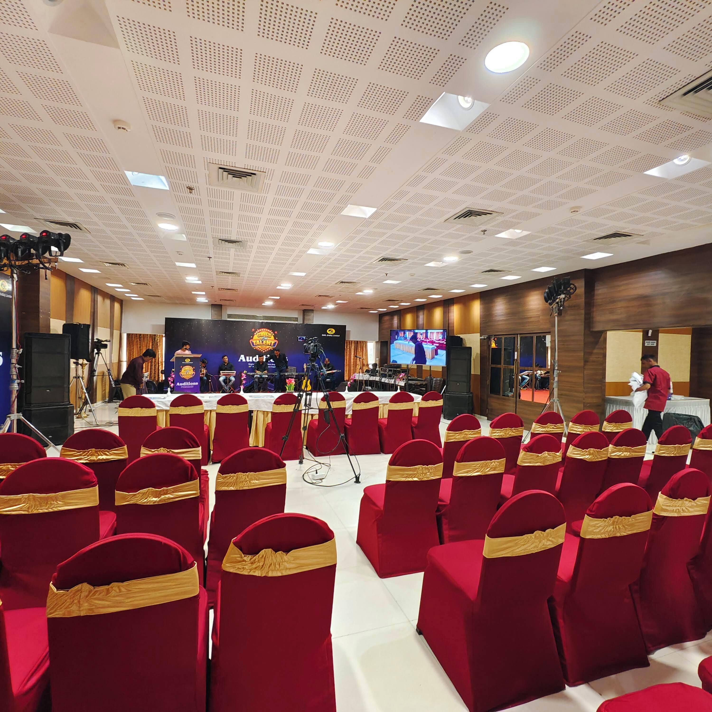 Un salón de conferencias con filas de sillas rojas con cintas doradas, un escenario con un logotipo y una pantalla grande.