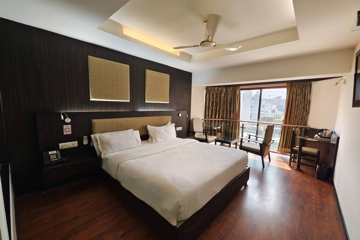 A modern hotel room with a large bed, a desk, and a balcony with a view of the city.