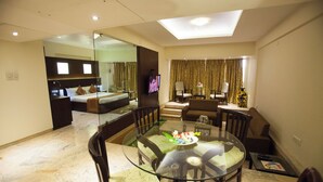 A modern hotel room with a glass dining table, comfortable seating, a large bed, a television, and a balcony with curtains. - The Oasis -Vadodara  (Vadodara)