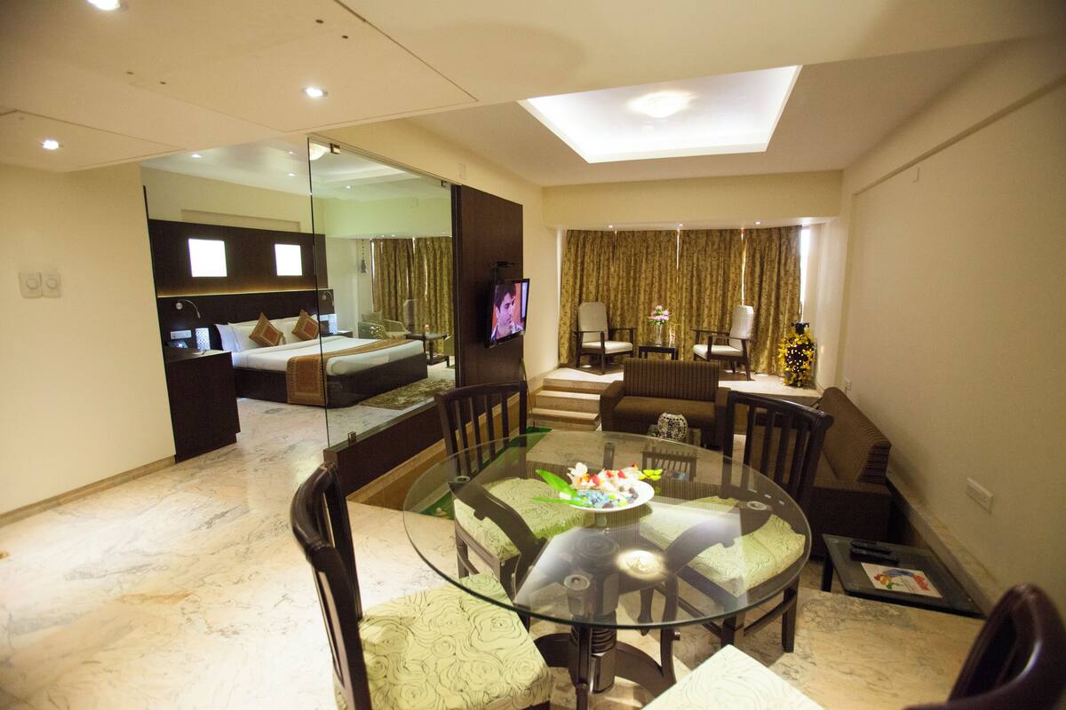 A modern hotel room with a glass dining table, comfortable seating, a large bed, a television, and a balcony with curtains.