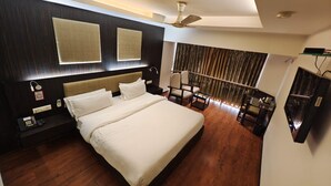 A modern hotel room with a large bed, a ceiling fan, a desk with a chair, a minibar, and a view of the city through the window. - The Oasis -Vadodara  (Vadodara)